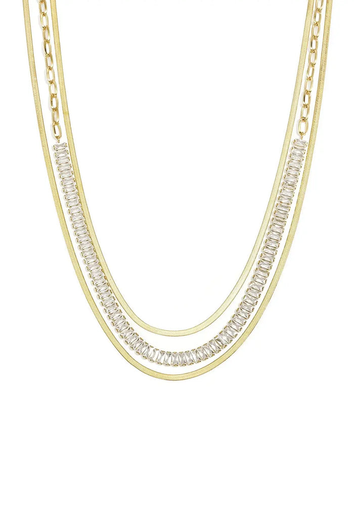 A gold-plated stainless steel multi-layered necklace featuring two flat herringbone chains framing a paperclip link chain with channel-set baguette cubic zirconia crystals against a white background. celeste-layered-herringbone-baguette-crystal-necklace.webp