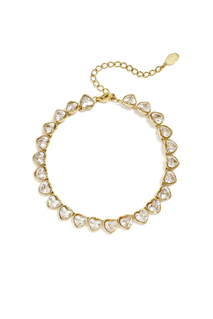 A gold-plated stainless steel tennis bracelet featuring a continuous line of heart-cut clear cubic zirconia crystals with a lobster clasp and extender chain against a white background. esme-heart-cut-crystal-tennis-bracelet.webp