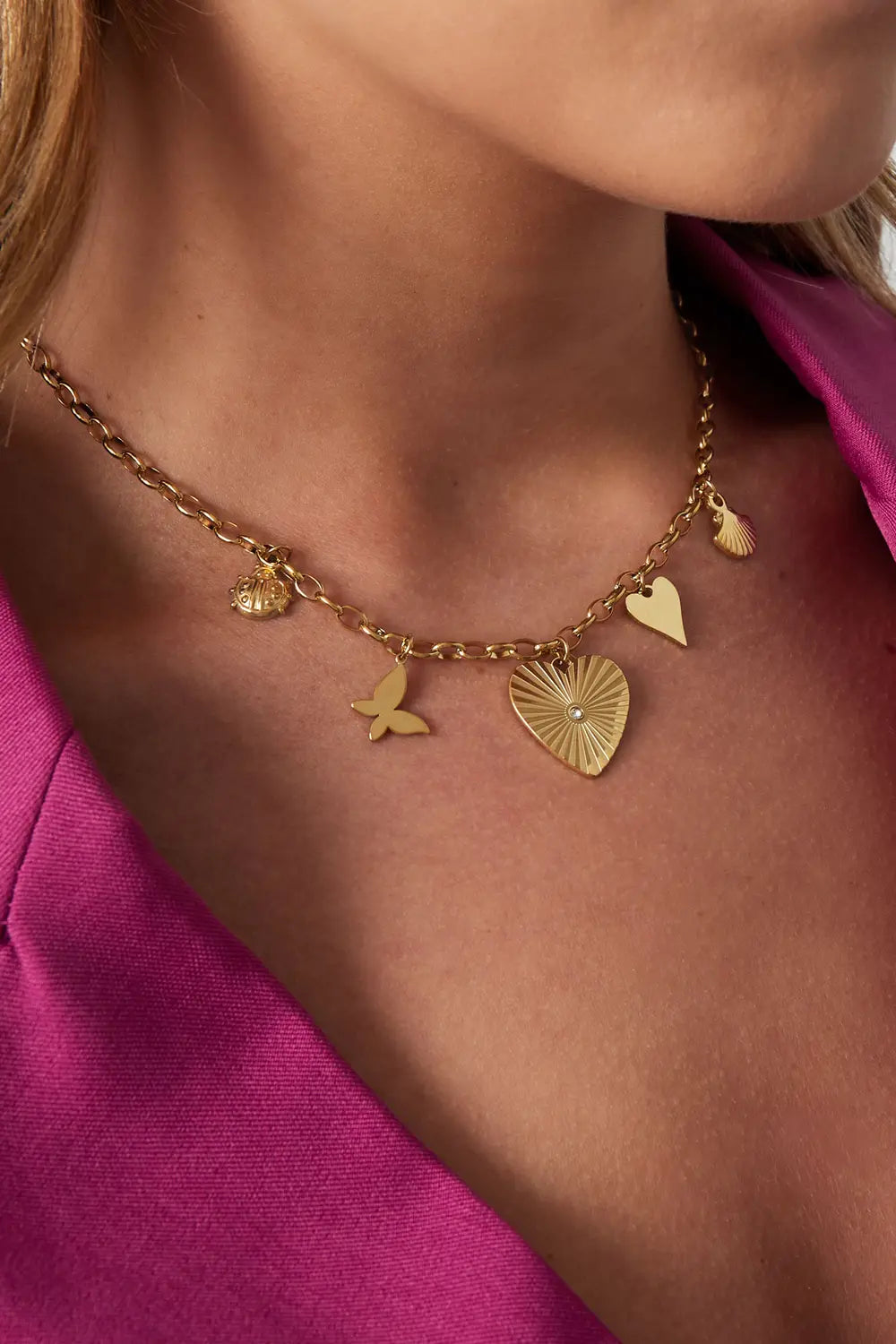 A gold-plated charm necklace featuring a central engraved sunburst heart pendant with a center crystal, paired with dangling charms of a ladybug, a butterfly, a small heart, and a scallop shell. meridien-sunburst-heart-charm-necklace.webp