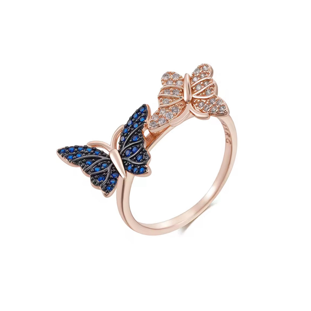 A 585 plated rose gold bypass ring featuring two detailed butterflies, one encrusted with sapphire blue crystals and the other with clear white crystals, on a white background.