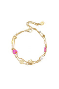 A gold-plated stainless steel double-layer charm bracelet featuring a central brushed gold flower, a hammered coin, a heart station, a white pearl, and a mix of magenta and yellow glass beads against a white background. posie-layered-eclectic-charm-bracelet.webp