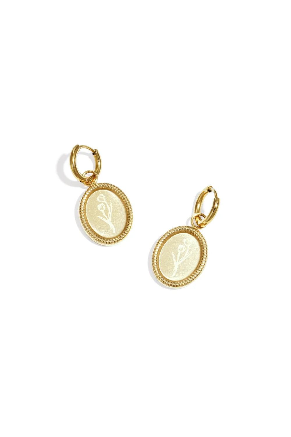 A pair of gold-plated stainless steel huggie hoop earrings featuring a dangling oval medallion coin with a detailed rope-textured border and a delicate engraved tulip flower sprig against a white background. tulipa-oval-floral-coin-huggie-earrings.webp