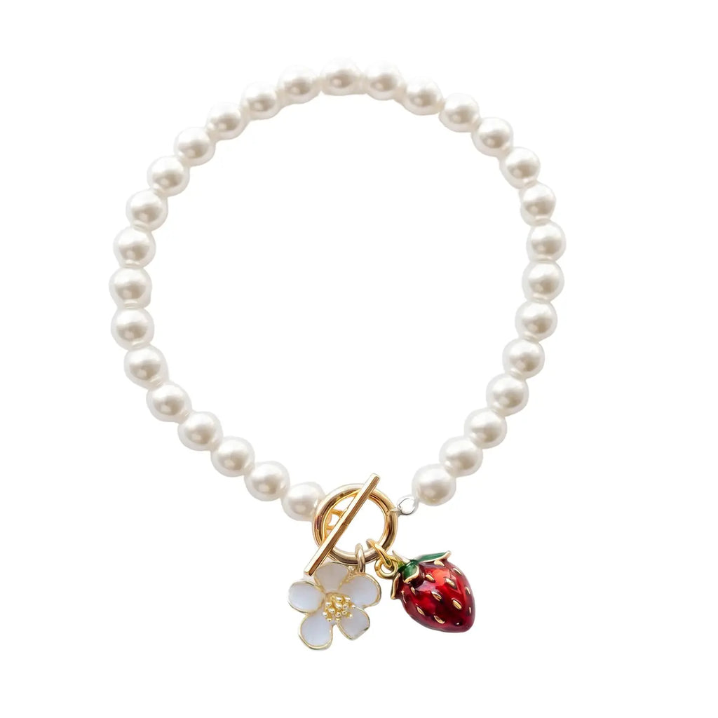 A pearl strand bracelet by Bill Skinner featuring a gold-plated front toggle clasp suspending a hand-painted red enamel strawberry charm and a white blossom charm. bill-skinner-strawberry-pearl-bracelet.webp