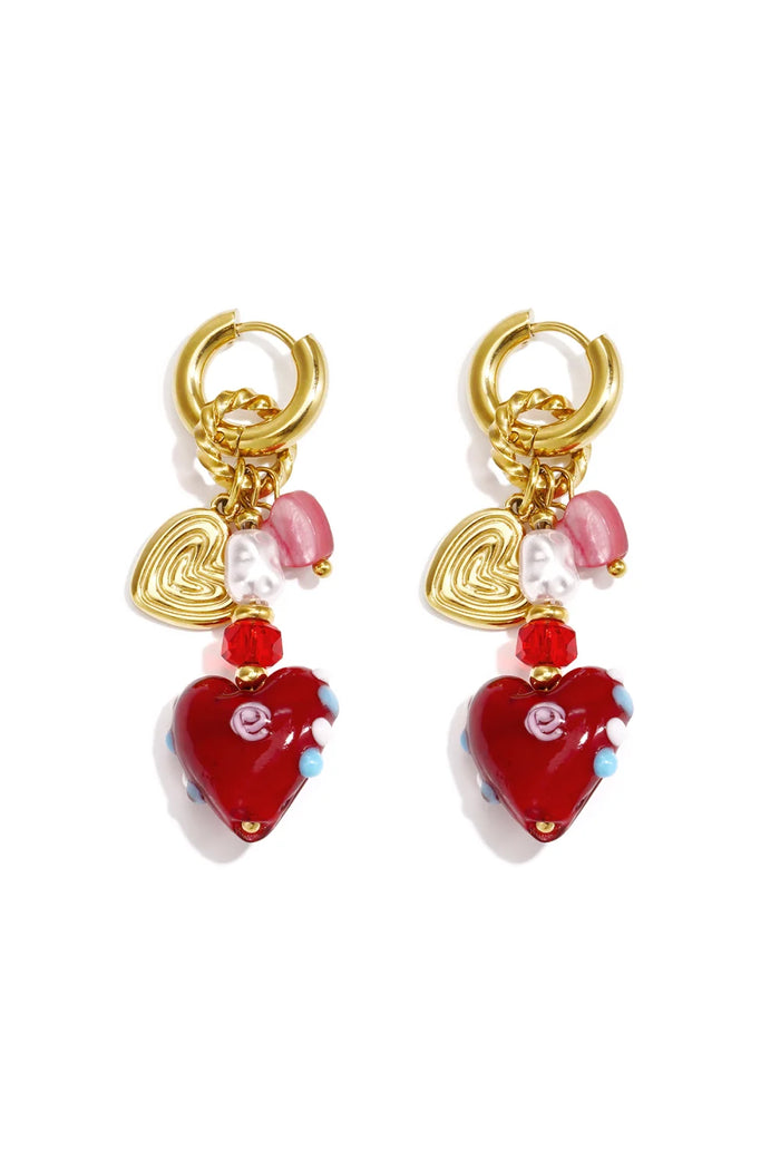 A pair of gold huggie hoop earrings with a layered charm cluster featuring a ridged gold heart, baroque pearl, blush pink cat-eye bead, red crystal spacer, and a deep red lampwork glass heart pendant with hand-painted blue dots and pink swirl detail, on a white background.